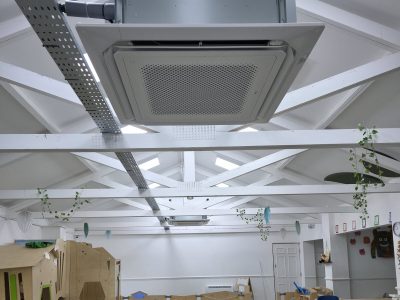 Air conditioning ceiling unit installed in bright room with decorative hanging plants and children’s play area.
